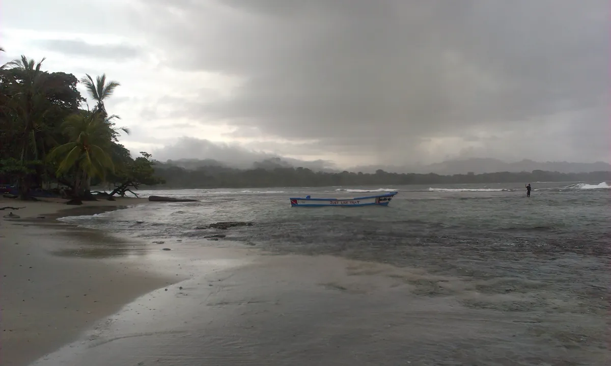 Overcast beach at Puerto Viejo de Talamanca with a small boat in shallow water near the shore, palm trees and hills in the background, photographed in 2015.